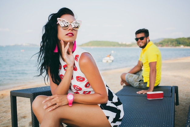 Schöne Frau des jungen stilvollen Hipsters, die am Strand sitzt, flirtend, sexy, heiß, Mode-Outfit, trendige Sonnenbrille, tropischer Urlaub, Urlaubsromantik, Honigmond, Mann auf Hintergrund schauend, lächelnd, glücklich