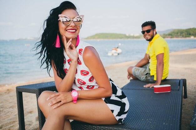 Schöne Frau des jungen stilvollen Hipsters, die am Strand sitzt, flirtend, sexy, heiß, Mode-Outfit, trendige Sonnenbrille, tropischer Urlaub, Urlaubsromantik, Honigmond, Mann auf Hintergrund schauend, lächelnd, glücklich