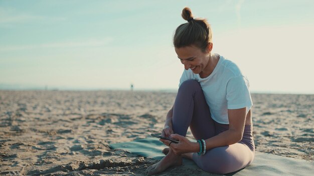 Schöne Frau, die auf der Matte sitzt und ihr Smartphone am Strand überprüft Junge Yogi-Frau sieht glücklich aus, wenn sie mit dem Handy am Meer ruht