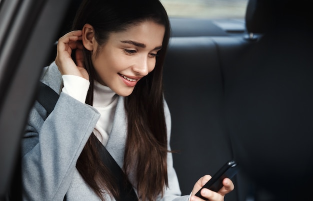 Schöne Frau, die auf Rücksitz sitzt und Textnachricht auf ihrem Handy liest, Sicherheitsgurt tragend, während im Auto unterwegs ist