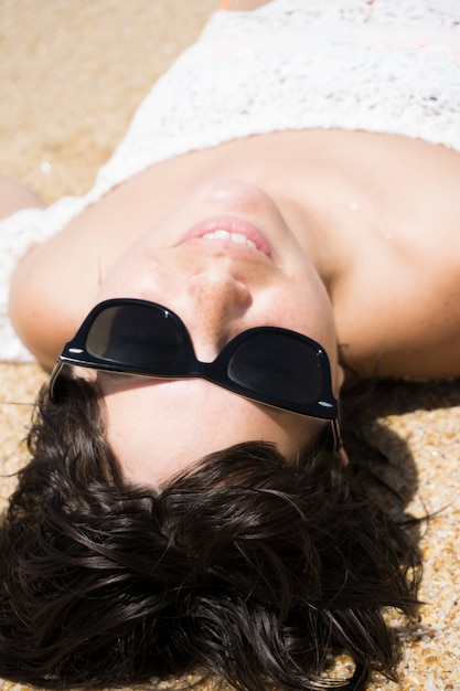 Schöne Frau, die auf Sand im weißen Kleid und in der Sonnenbrille während des Tages liegt.