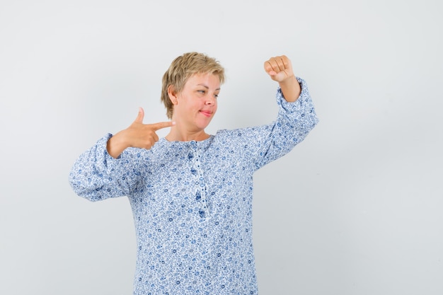 Kostenloses Foto schöne frau, die aufwirft, als würde sie etwas erhöhen, während sie in gemusterter bluse darauf zeigt und konzentriert aussieht. vorderansicht.