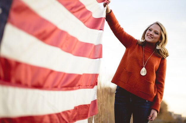 Schöne Frau, die die amerikanische Flagge hält, während sie nahe den Bäumen steht