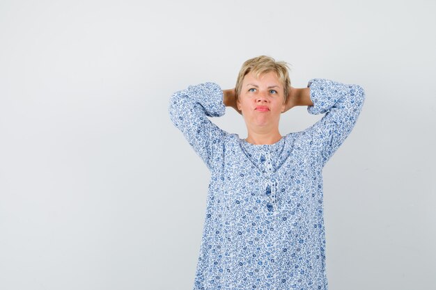 Schöne Frau, die Hände hinter Kopf in gemusterter Bluse hält und nachdenklich aussieht. Vorderansicht.