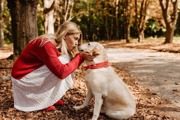 Schöne Frau, die ihren entzückenden schönen Hund küsst. Schönes Mädchen im roten Pullover und im weißen Kleid, die Liebe mit einem Haustier teilen.