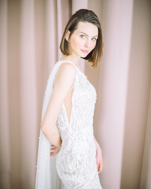 Schöne Frau, die im Raum mit Perlenhintergrund im langen weißen Kleid steht und schaut.