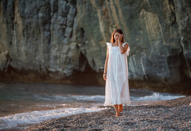 Schöne Frau, die im weißen Kleid an der Küste während des Sonnenuntergangs geht und traurig schaut.