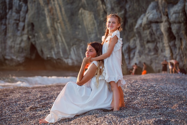 Schöne Frau, die im weißen Kleid mit ihrer Tochter an der Küste während des Sonnenuntergangs sitzt.