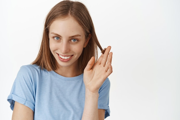 Schöne Frau, die kokett lächelt, mit Haarsträhne spielt und glücklich und süß nach vorne blickt, flirtet, im T-Shirt gegen weiße Wand steht.