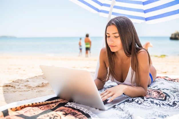 Schöne frau, die online auf laptop arbeitet, während am strand unter sonnenschirm nahe meer liegt. glückliches lächelndes freiberufliches mädchen, das sich entspannt und notebook für freiberufliche internetarbeit verwendet.