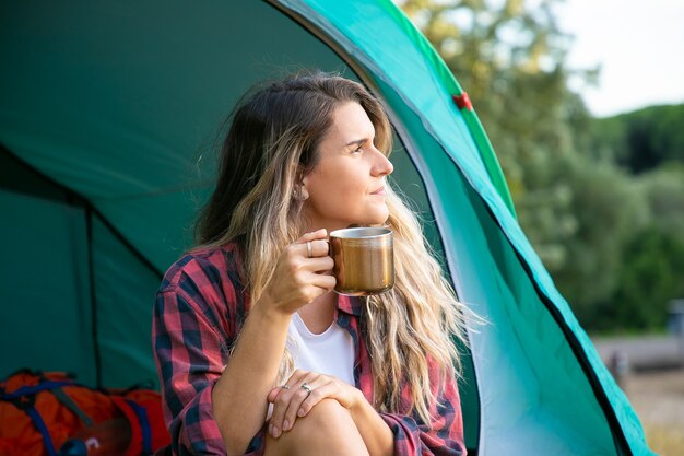 Schöne Frau, die Tasse Tee hält, im Zelt sitzt und wegschaut. Kaukasische Wanderin, die sich auf Natur entspannt, genießt und kampiert. Backpacking Tourismus, Abenteuer und Sommerurlaub Konzept