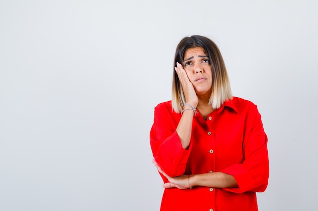 Schöne Frau, die Wange auf der Handfläche lehnt, in roter Bluse aufschaut und verzweifelt aussieht, Vorderansicht.