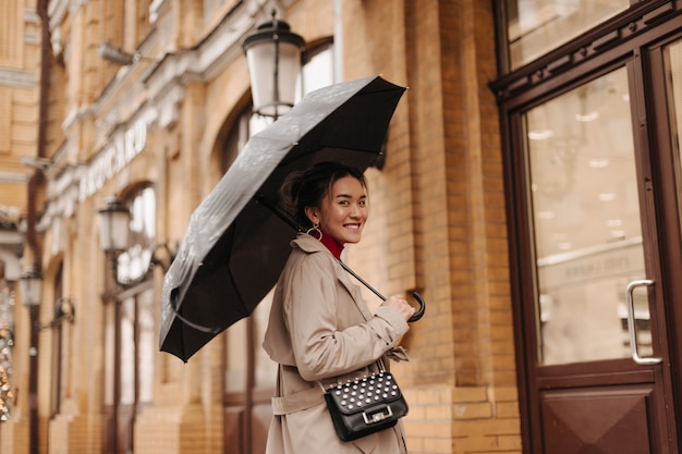 Schöne Frau im beige Trenchcoat mit Umhängetasche mit Lächeln geht unter Regenschirm in der europäischen Stadt.