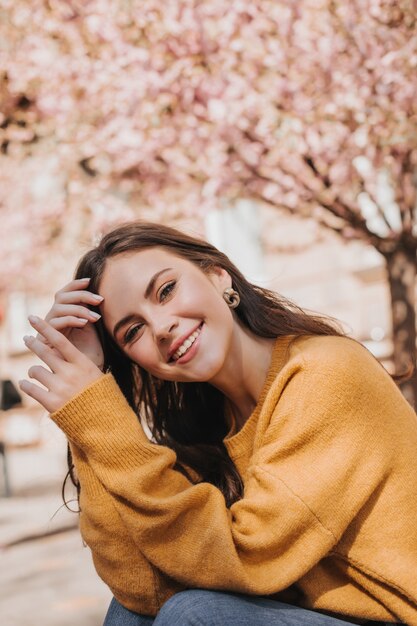 Schöne Frau im hellen Pullover, der auf Straße gegen Hintergrund von Sakura aufwirft. Stadtporträt der attraktiven Dame im gelben Outfit breit lächelnd