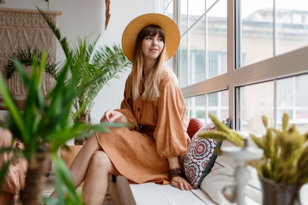 Schöne Frau im Leinenkleid und im Strohhut, die in der Wohnung im Boho-Stil aufwirft