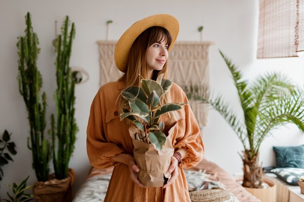 Schöne Frau im Leinenkleid und im Strohhut, die in der Wohnung im Boho-Stil aufwirft