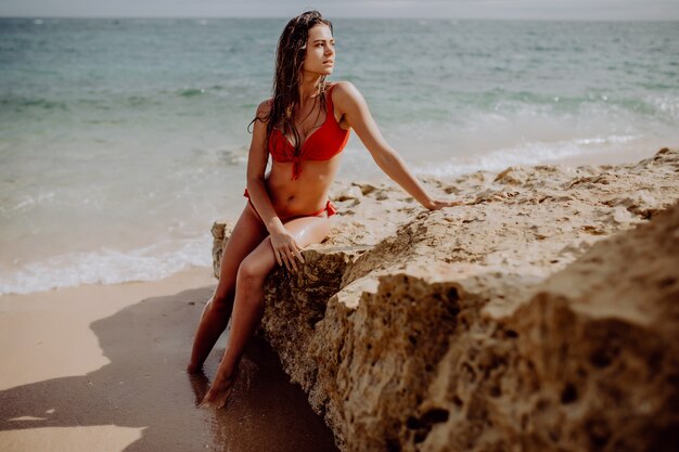 Schöne Frau im roten Bikini, der am Strand auf Felsen sitzend aufwirft