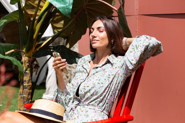 Schöne Frau im Sommerkleid sitzt am sonnigen Tag mit Handy auf Stuhl im Hinterhof