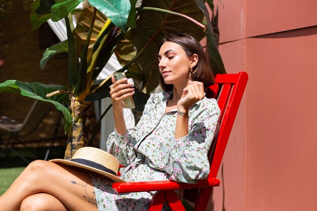 Schöne Frau im Sommerkleid sitzt am sonnigen Tag mit Handy auf Stuhl im Hinterhof
