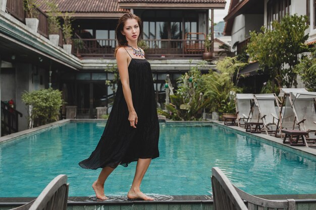 Schöne Frau im stilvollen langen schwarzen Kleid, das am Pool der tropischen Villa, eleganter Sommerstil, Urlaub, Modetrend, barfuß gehen aufwirft