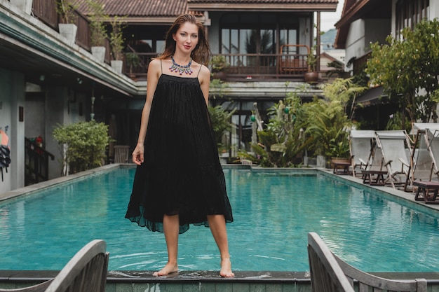 Schöne Frau im stilvollen langen schwarzen Kleid, das am Pool der tropischen Villa, eleganter Sommerstil, Urlaub, Modetrend, barfuß gehen aufwirft