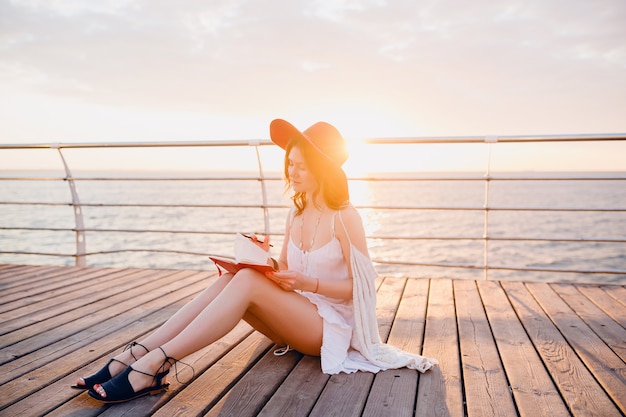 Schöne frau im weißen kleid, das am meer auf sonnenaufgang denkt und notizen im tagebuchbuch macht