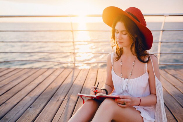 Schöne Frau im weißen Kleid, das am Meer auf Sonnenaufgang denkt und Notizen im Tagebuchbuch macht