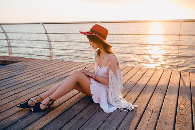 Schöne Frau im weißen Kleid, das am Meer auf Sonnenaufgang denkt und Notizen im Tagebuchbuch macht