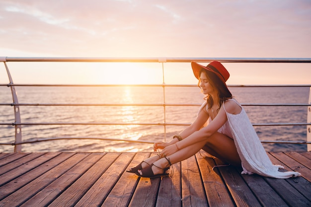 Schöne Frau im weißen Kleid, das am Meer auf Sonnenaufgang in der romantischen Stimmung sitzt