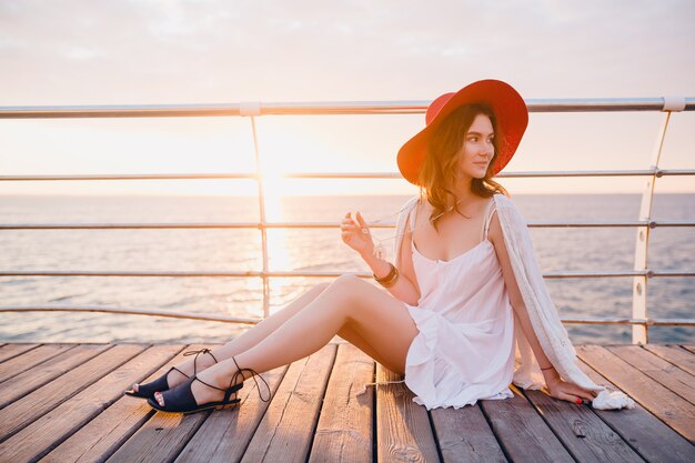 Schöne Frau im weißen Kleid sitzt am Meer auf Sonnenaufgang in der romantischen Stimmung, die roten Hut trägt