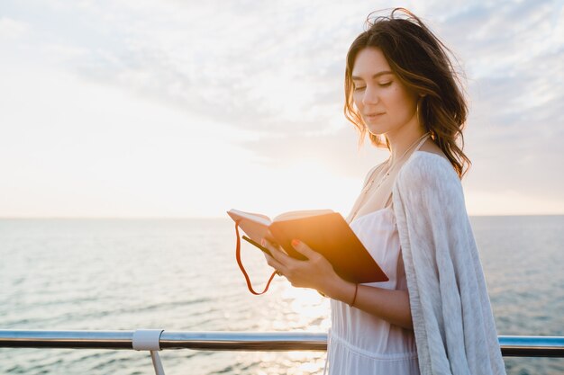 Schöne Frau im weißen Sommerkleid, das durch das Meer auf Sonnenaufgang mit Tagebuchbuch in der romantischen Stimmung denkt und Notizen macht