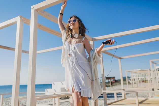 Schöne Frau im weißen Sommerkleid, das Musik auf Kopfhörern hört, die tanzen und Spaß haben, Smartphone halten, Sommerferienart-Seestrand