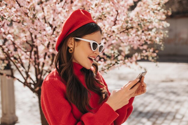 Schöne Frau in Baskenmütze und Sonnenbrille plaudert am Telefon in der Nähe von Sakura. Außenporträt der Dame im roten Kaschmirpullover, der Handy hält