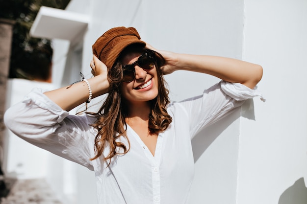 Schöne Frau in der Sonnenbrille trägt Cordmütze. Porträt des Mädchens in Hochstimmung gegen weiße Wand.