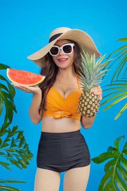 Schöne Frau in einem Badeanzug, der Ananas und Wassermelone auf Blau hält