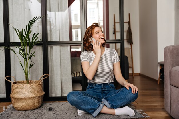 Schöne Frau in Jeans sitzt auf dem Boden im Wohnzimmer und telefoniert.