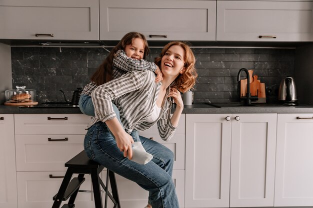 Schöne Frau in Jeans sitzt auf Stuhl in der Küche, während ihre Tochter sie von hinten umarmt.