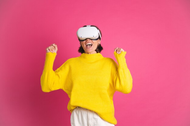 Schöne Frau in leuchtend gelbem Pullover auf Rosa in Virtual-Reality-Brille glücklich springende Faustsiegergeste