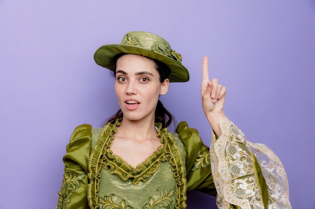 Schöne Frau in Renaissance-Kleid und Hut mit einem Lächeln auf einem intelligenten Gesicht, das mit dem Zeigefinger nach oben zeigt und eine neue Idee auf Blau hat