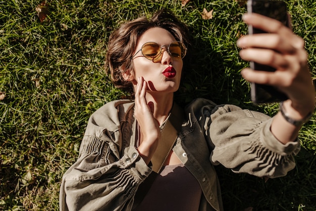 Schöne Frau in runder Sonnenbrille und Jacke liegt auf Gras und bläst Kuss draußen. Junge Frau mit kurzen Haaren, die selfie im Freien nehmen.