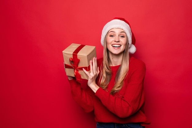 Schöne Frau in Weihnachtsmütze mit Geschenkbox in Händen isoliert auf roter Wand