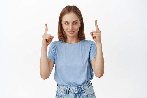 Schöne Frau mit blonden Haaren und blauen Augen, die mit den Fingern auf den leeren Raum zeigt, das Verkaufsrabattbanner zeigt, auf den Link klickt, glücklich lächelt und gegen die weiße Wand steht.