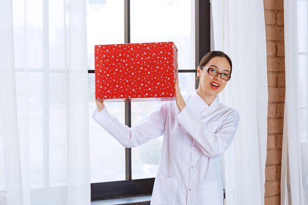 Schöne Frau mit Brille im Laborkittel, die große Geschenkbox in der Nähe des Fensters hält.