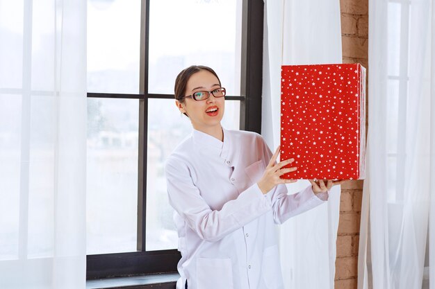 Schöne Frau mit Brille im Laborkittel, die große Geschenkbox in der Nähe des Fensters hält.