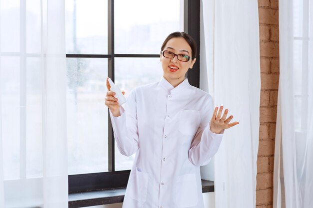 Schöne Frau mit Brille im Laborkittel, die Sprühdesinfektionsmittel in der Nähe des Fensters hält.