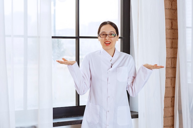 Schöne Frau mit Brille und Laborkittel, die in der Nähe des Fensters steht und die Schultern zuckt.