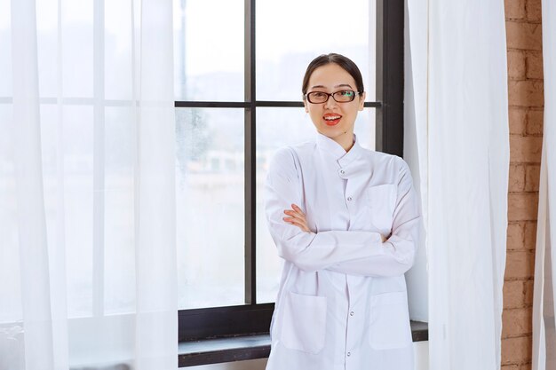 Schöne Frau mit Brille und Laborkittel, die mit verschränkten Armen in der Nähe des Fensters steht.