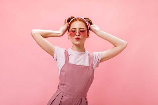 Schöne Frau mit Brötchen gekleidet in rosa Overalls und weißem T-Shirt, das mit geschlossenen Augen auf rosa Hintergrund aufwirft.