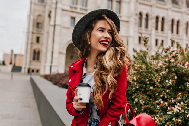 Schöne Frau mit eleganter gewellter Frisur, die weg schaut, während Kaffee auf der Straße trinkt
