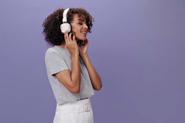 Schöne Frau mit Kopfhörern, die Musik auf lila Hintergrund genießt Charmantes Mädchen in grauem T-Shirt und weißem Rock, das ihre Lieblingslieder hört